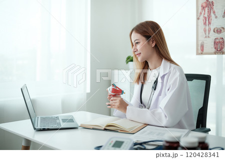 A woman in a white lab coat is sitting at a desk with a laptop 120428341