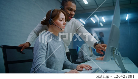 Female hotline operator in headset monitors data on computer with African American colleague Female hotline operator in headset monitors data on computer with African American colleague 120428891