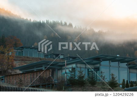 Luxury houses in autumn mountains covered with clouds in the morning. Modern hotel in Carpathian mountains, Ukraine. Foggy morning in mountain resort. House in autumn forest on foggy hills.  120429226