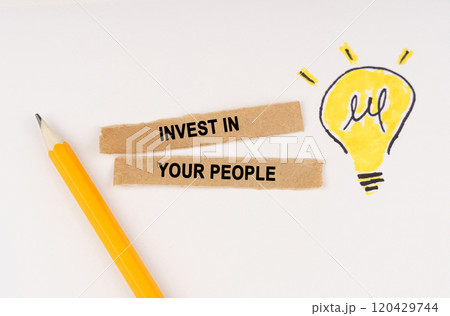 On a white surface, a luminous light bulb and pieces of paper with the inscription - Invest in Your People 120429744
