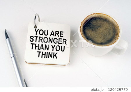 On a white surface, a cup of coffee, a pen and a notepad with the inscription - You Are Stronger Than You Think 120429779