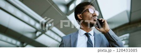 A professional businessman in a suit standing outside and talking on smartphone, reflecting light from his glasses while looking ahead, steel architectural details in background 120431085