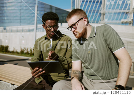 Two young men sitting outdoors, discussing project on a tablet. Background includes modern architectural elements and clear sky, suggesting a productive, open environment 120431110