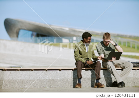 Two men sitting on bench are discussing work using digital tablet. Modern architectural building in background creates dynamic and futuristic atmosphere enhancing conversation 120431111