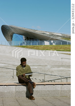 Man sitting on concrete steps while reading book outdoors near a uniquely designed modern building with interesting architectural elements visible in background 120431112