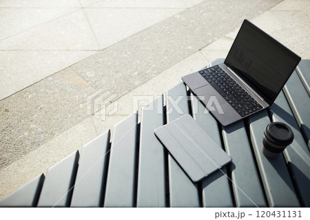Laptop with open screen, coffee mug, and case on outdoor bench. Bench and pavement creating urban, work-friendly environment under daylight Laptop with open screen, coffee mug, and case on outdoor bench. Bench and pavement creating urban, work-friendly environment under daylight 120431131