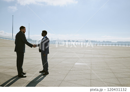 Two business professionals shaking hands standing in a spacious outdoor area surrounded by large cityscape on horizon. Background features a vast, open space under clear blue sky Two business professionals shaking hands standing in a spacious outdoor area surrounded by large cityscape on horizon. Background features a vast, open space under clear blue sky 120431132