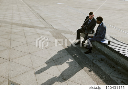 Two businessmen engaged in discussion while sitting on a bench in an outdoor urban area, creating shadows on pavement, reflecting corporate attire and professional interaction 120431133