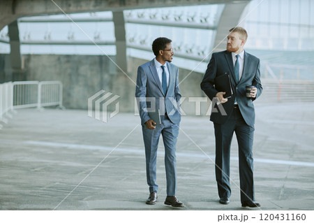 Two business colleagues dressed in formal suits walking outdoors in urban environment, engaged in conversation and holding coffee cup and documents 120431160