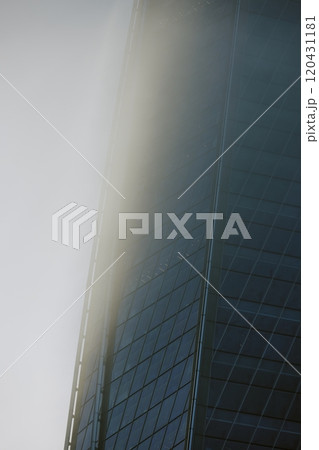 Tall skyscraper partially enveloped by thick fog in an urban setting, accentuating its sleek architectural design and futuristic appeal, with a predominantly glass facade 120431181