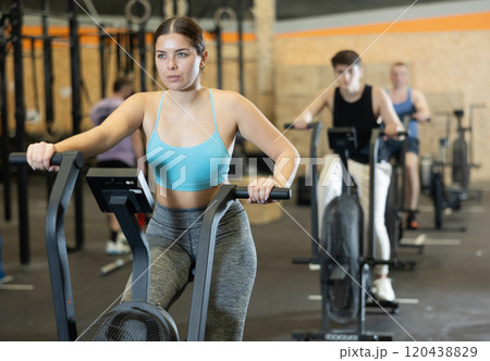 Young motivated fitness woman warming up and pedaling on exercise bike during group CrossFit workout in gym health center Young motivated fitness woman warming up and pedaling on exercise bike during group CrossFit workout in gym health center 120438829