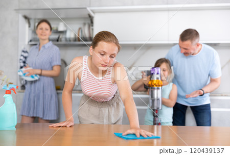 Bored teenage girl wiping table while cleaning apartment with family Bored teenage girl wiping table while cleaning apartment with family 120439137