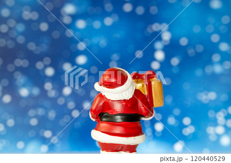 Cheerful Santa Claus figurine holding a golden gift box, set against a vibrant blue background with snowy bokeh effects Cheerful Santa Claus figurine holding a golden gift box, set against a vibrant blue background with snowy bokeh effects 120440529