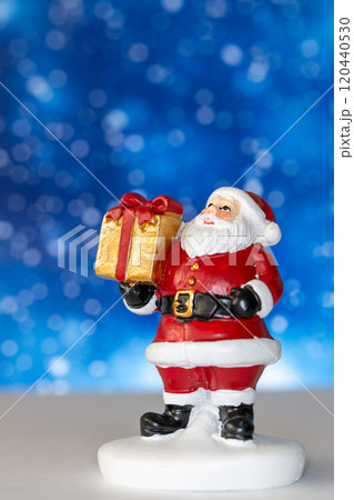 Cheerful Santa Claus figurine holding a golden gift box, set against a vibrant blue background with snowy bokeh effects Cheerful Santa Claus figurine holding a golden gift box, set against a vibrant blue background with snowy bokeh effects 120440530