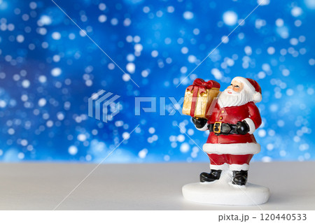 Cheerful Santa Claus figurine holding a golden gift box, set against a vibrant blue background with snowy bokeh effects Cheerful Santa Claus figurine holding a golden gift box, set against a vibrant blue background with snowy bokeh effects 120440533