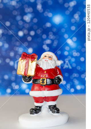 Cheerful Santa Claus figurine holding a golden gift box, set against a vibrant blue background with snowy bokeh effects Cheerful Santa Claus figurine holding a golden gift box, set against a vibrant blue background with snowy bokeh effects 120440538