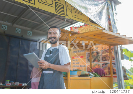 A joyful vendor serves customers at a colorful stall, using a tablet to enhance service 120441727