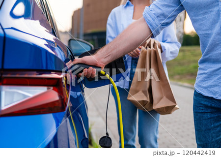 Person charges an electric car while another holds shopping bags at an urban parking lot 120442419