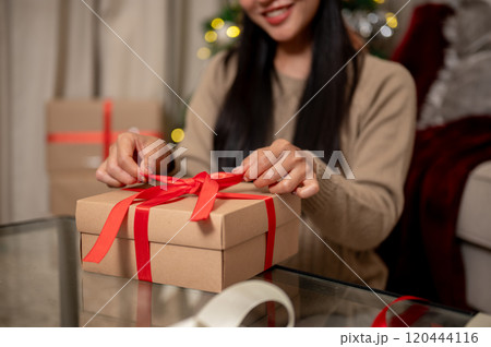 A close-up of a happy woman tying a red ribbon around a Christmas gift box in the living room. 120444116