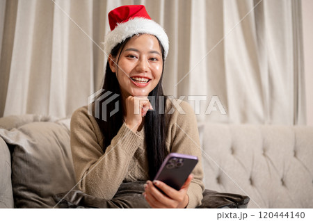 A beautiful Asian woman in cozy clothes and a Christmas hat sits on the sofa in the living room. A beautiful Asian woman in cozy clothes and a Christmas hat sits on the sofa in the living room. 120444140
