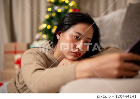 A sad, lonely Asian woman sits on the sofa, feeling bored, celebrating Christmas night alone at home A sad, lonely Asian woman sits on the sofa, feeling bored, celebrating Christmas night alone at home 120444141