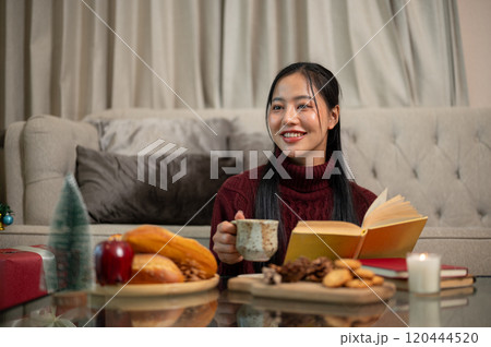 A happy woman sits in her living room, enjoying a book and some snacks during the Christmas holiday. A happy woman sits in her living room, enjoying a book and some snacks during the Christmas holiday. 120444520