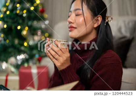 A calm Asian woman in a cozy red sweater enjoys a hot drink, spending the Christmas at home alone. A calm Asian woman in a cozy red sweater enjoys a hot drink, spending the Christmas at home alone. 120444523