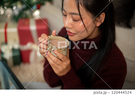 A calm Asian woman in a cozy red sweater enjoys a hot drink, spending the Christmas at home alone. A calm Asian woman in a cozy red sweater enjoys a hot drink, spending the Christmas at home alone. 120444524