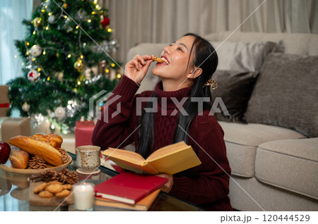 A cheerful Asian woman enjoys Christmas cookies and a warm drink while reading in her living room. 120444529