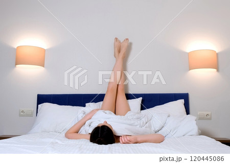 Brunette woman is resting lying in bed with legs on a wall in hotel room. Brunette woman is resting lying in bed with legs on a wall in hotel room. 120445086