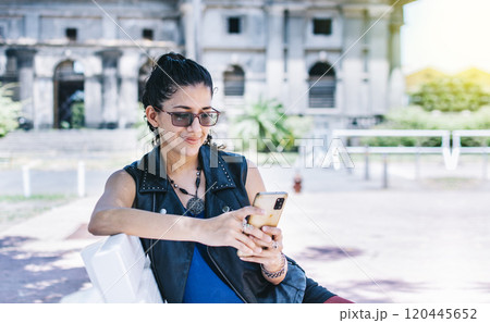 Urban style girl sitting on a bench with her cell phone, An attractive girl sitting on the cell phone smiling, Girl sitting on a bench texting on her cell phone. 120445652