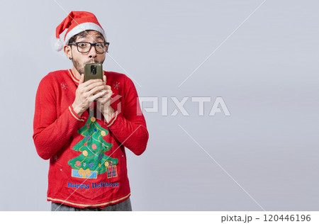 Man in christmas hat with amazed face holding phone. Online Christmas special offers concept. Young man in christmas clothes and hat amazed holding the phone 120446196