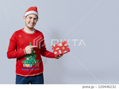 Smiling guy in christmas hat holding and pointing a gift box. Portrait of teenager in christmas hat holding and pointing a gift box. Guy in christmas hat and sweater holding a gift box isolated 120446232