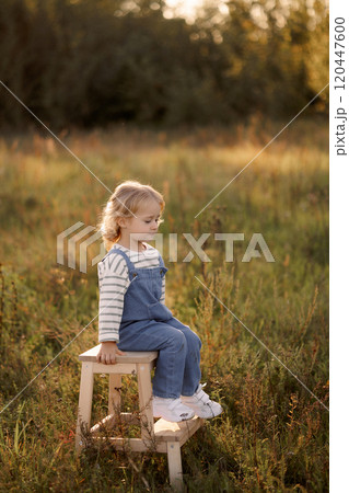 Little smiling child in denim overalls sits on a wooden stool in the field.  120447600