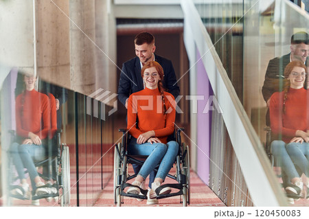 A company director assists his business colleague in a wheelchair, helping her navigate to their startup office, where they work alongside their diverse team of colleagues, emphasizing inclusivity and A company director assists his business colleague in a wheelchair, helping her navigate to their startup office, where they work alongside their diverse team of colleagues, emphasizing inclusivity and 120450083