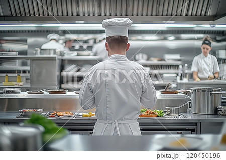 A chef in a white uniform stands in a modern kitchen, rear view, and prepares food, kitchen equipment in the background 120450196
