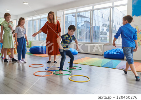 Small nursery school children with female teacher on floor indoors in classroom, doing exercise. Jumping over hula hoop circles track on the floor. Small nursery school children with female teacher on floor indoors in classroom, doing exercise. Jumping over hula hoop circles track on the floor. 120450276