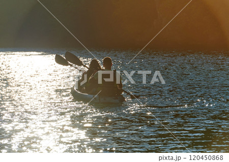 A young couple enjoying an idyllic kayak ride in the middle of a beautiful river surrounded by forest greenery in sunset time  120450868
