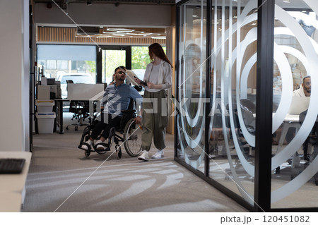 Young business colleagues, collaborative business colleagues, including a person in a wheelchair, walk past a modern glass office corridor, illustrating diversity, teamwork and empowerment in the 120451082