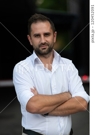 A successful businessman in a white shirt, with crossed arms, poses outdoors, confident expression on his face. A successful businessman in a white shirt, with crossed arms, poses outdoors, confident expression on his face. 120452091