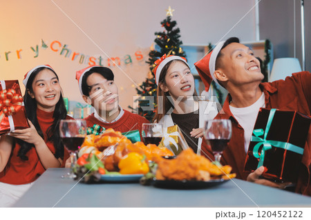 Group of young Asian man and women as friends having fun at a New Year's celebration, holding gift boxes standing by Christmas tree decoration, midnight countdown Party at home with holiday season. 120452122