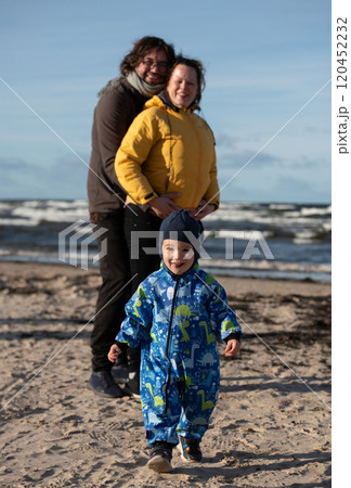 A happy family strolls along a chilly autumn beach, with their young son joyfully running ahead, creating lasting memories as the crisp wind and soothing waves embrace them in a moment of pure 120452232