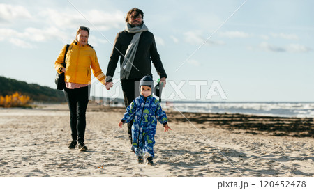 A happy family strolls along a chilly autumn beach, with their young son joyfully running ahead, creating lasting memories as the crisp wind and soothing waves embrace them in a moment of pure A happy family strolls along a chilly autumn beach, with their young son joyfully running ahead, creating lasting memories as the crisp wind and soothing waves embrace them in a moment of pure 120452478