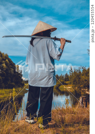 Modern samurai in a conical hat stands by the river holding a katana, lost in the tranquility of nature. Beautiful scene with a samurai holding a katana on his shoulder on the bank of a river. 120455554