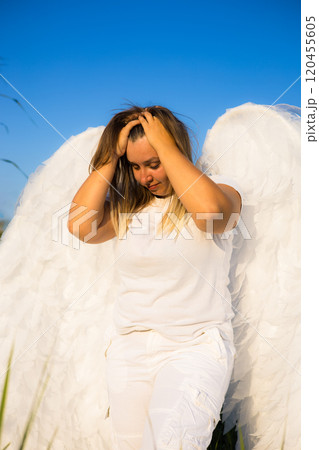 Angel upset and sad. Young woman with big white angel wings posing in a field with her hands in her hair under a clear blue summer sky, creating a serene and airy atmosphere. Angel upset and sad. Young woman with big white angel wings posing in a field with her hands in her hair under a clear blue summer sky, creating a serene and airy atmosphere. 120455605