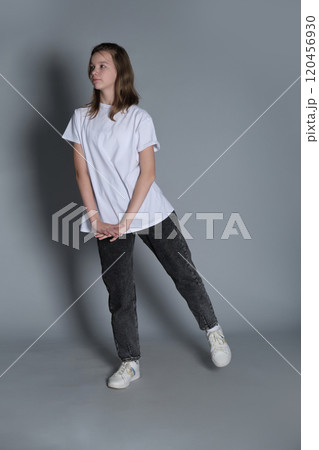 A young girl leans slightly to one side, her hands gently clasped, dressed in casual jeans and a white t-shirt. The minimalist background emphasizes her calm and carefree expression. 120456930