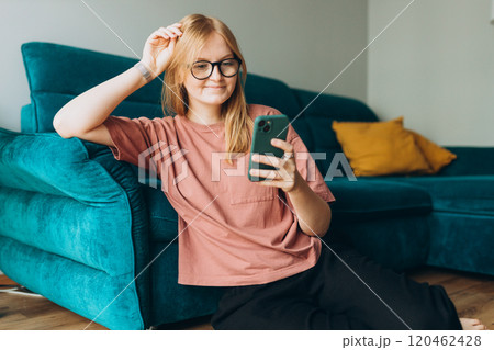 Beautiful Caucasian pleased woman smiling and using cellphone while sitting near sofa on floor in apartment. Phone Communication and comfort concept 120462428