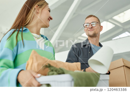 Medium shot of smiling man looking at cheerful girlfriend carrying boxes with personal belongings to self storage warehouse, while donating items to charity or moving out 120463386