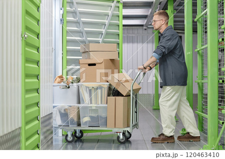 Full length shot of handsome man loading cart with cardboard boxes into self storage unit at warehouse while moving out or decluttering apartment 120463410