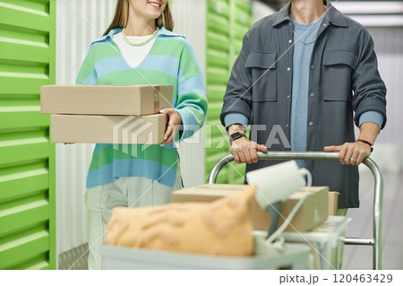 Cropped shot of smiling woman carrying pile of cardboard boxes walking next to boyfriend helping with moving out, while pushing loaded dolly cart in hallway of self storage warehouse 120463429